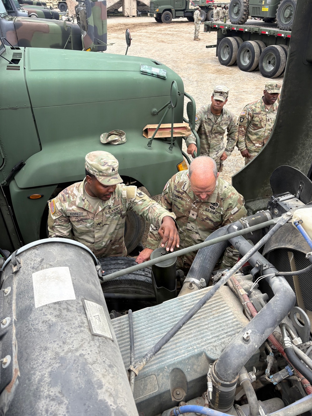 U.S. Army soldiers are inspecting or performing maintenance on the engine compartment of a military tactical vehicle. Two soldiers in the foreground — name tapes visible reading 'KENNEDY' and 'THOMAS' — are leaning over an open engine bay, while two additional soldiers observe from behind. The setting appears to be a military motor pool or field staging area with multiple military vehicles visible in the background.