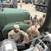 U.S. Army soldiers are inspecting or performing maintenance on the engine compartment of a military tactical vehicle. Two soldiers in the foreground — name tapes visible reading 'KENNEDY' and 'THOMAS' — are leaning over an open engine bay, while two additional soldiers observe from behind. The setting appears to be a military motor pool or field staging area with multiple military vehicles visible in the background.