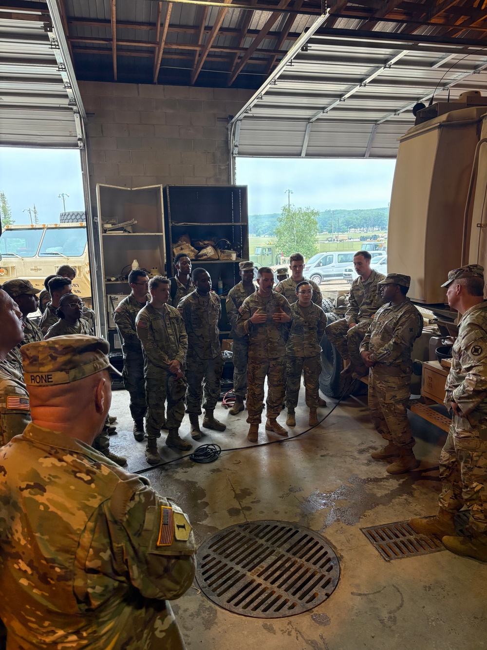 A group of U.S. Army soldiers in OCP (Operational Camouflage Pattern) uniforms are gathered inside a military maintenance bay or motor pool facility. One soldier appears to be briefing or addressing the group in a semi-circular formation. The setting includes military vehicles, tool storage units, and an open bay door revealing an outdoor motor pool area.