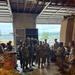 A group of U.S. Army soldiers in OCP (Operational Camouflage Pattern) uniforms are gathered inside a military maintenance bay or motor pool facility. One soldier appears to be briefing or addressing the group in a semi-circular formation. The setting includes military vehicles, tool storage units, and an open bay door revealing an outdoor motor pool area.