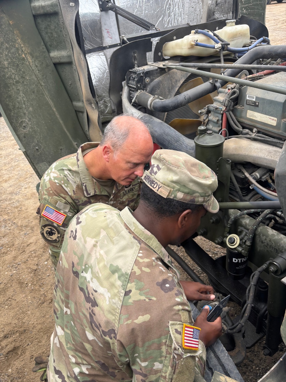Two U.S. Army soldiers in MultiCam/OCP camouflage uniforms are performing maintenance or inspection on a military vehicle engine. One soldier, identified by name tape as 'KENNEDY,' is in the foreground working directly on the engine components, while the other soldier leans in to observe or assist. Both wear American flag patches on their left shoulders, indicating active deployment or operational status.