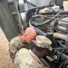 Two U.S. Army soldiers in MultiCam/OCP camouflage uniforms are performing maintenance or inspection on a military vehicle engine. One soldier, identified by name tape as 'KENNEDY,' is in the foreground working directly on the engine components, while the other soldier leans in to observe or assist. Both wear American flag patches on their left shoulders, indicating active deployment or operational status.