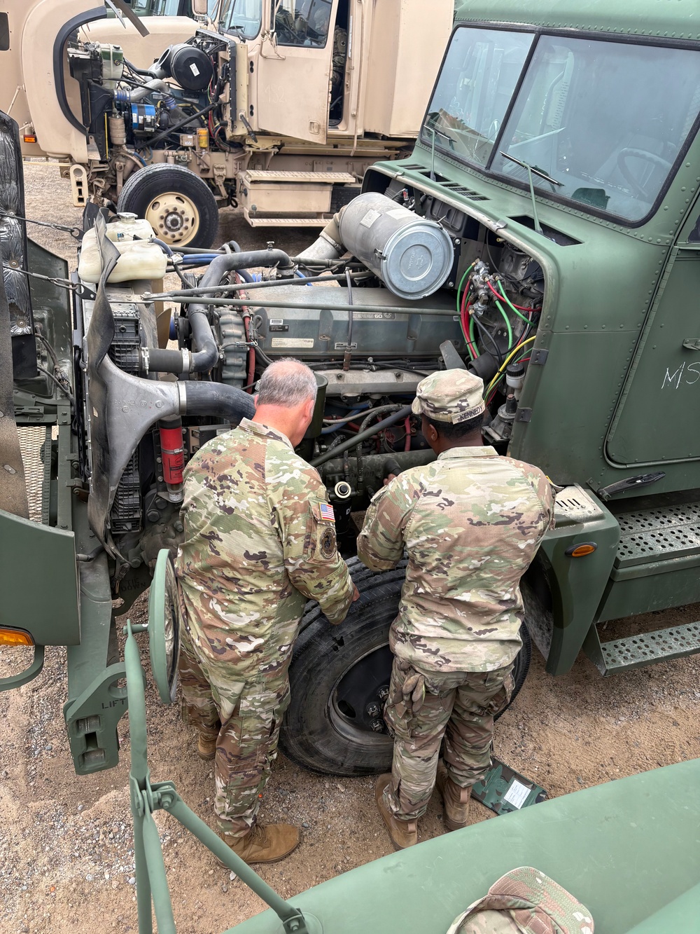 Two soldiers in US Army OCP (Operational Camouflage Pattern) uniforms are performing maintenance on a military tactical vehicle with its hood open, exposing the engine compartment. They are working in what appears to be a motor pool or field maintenance area surrounded by multiple military vehicles. A spare tire is visible on the ground between them.