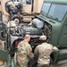 Two soldiers in US Army OCP (Operational Camouflage Pattern) uniforms are performing maintenance on a military tactical vehicle with its hood open, exposing the engine compartment. They are working in what appears to be a motor pool or field maintenance area surrounded by multiple military vehicles. A spare tire is visible on the ground between them.