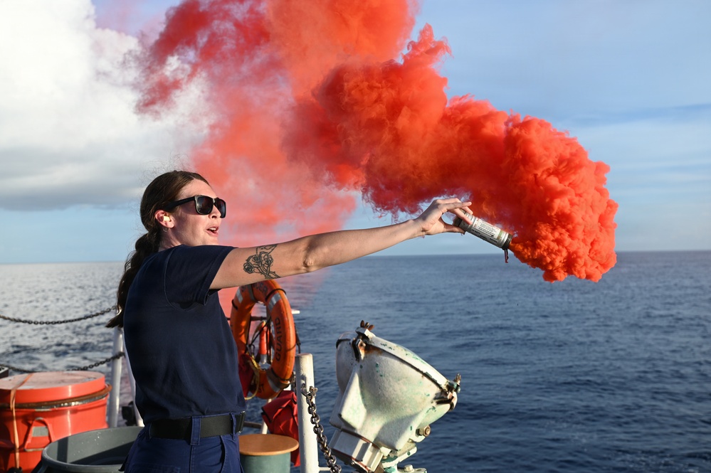 A woman in a dark navy uniform is standing on the deck of a vessel at sea, holding and discharging a handheld orange/red smoke signal flare. The bright red-orange smoke billows dramatically to the right against a partly cloudy blue sky. This appears to be a maritime safety drill or training exercise conducted aboard a Coast Guard or similar naval vessel.