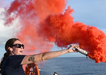 A woman in a dark navy uniform is standing on the deck of a vessel at sea, holding and discharging a handheld orange/red smoke signal flare. The bright red-orange smoke billows dramatically to the right against a partly cloudy blue sky. This appears to be a maritime safety drill or training exercise conducted aboard a Coast Guard or similar naval vessel.