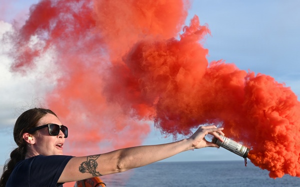 A woman in a dark navy uniform is standing on the deck of a vessel at sea, holding and discharging a handheld orange/red smoke signal flare. The bright red-orange smoke billows dramatically to the right against a partly cloudy blue sky. This appears to be a maritime safety drill or training exercise conducted aboard a Coast Guard or similar naval vessel.