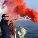 A woman in a dark navy uniform is standing on the deck of a vessel at sea, holding and discharging a handheld orange/red smoke signal flare. The bright red-orange smoke billows dramatically to the right against a partly cloudy blue sky. This appears to be a maritime safety drill or training exercise conducted aboard a Coast Guard or similar naval vessel.