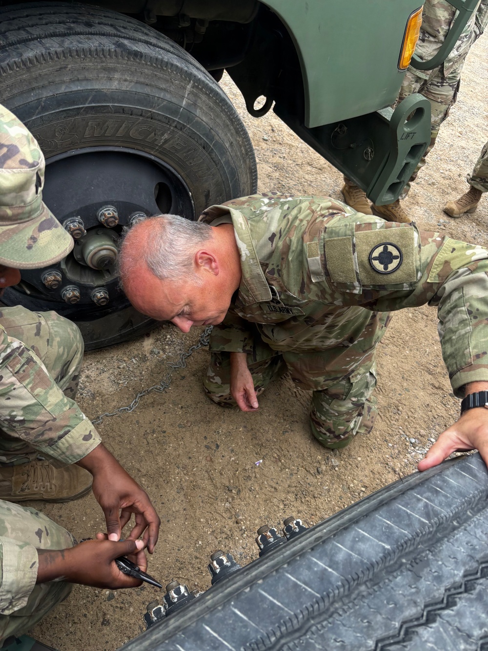 Multiple U.S. Army soldiers in OCP (Operational Camouflage Pattern) uniforms are working on or inspecting the undercarriage and wheel area of a military vehicle. The central figure, an older soldier with gray hair, is kneeling and leaning down to examine something beneath the vehicle. Other soldiers are assisting, with one appearing to mark or handle lug nuts/hardware on the ground.