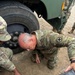 Multiple U.S. Army soldiers in OCP (Operational Camouflage Pattern) uniforms are working on or inspecting the undercarriage and wheel area of a military vehicle. The central figure, an older soldier with gray hair, is kneeling and leaning down to examine something beneath the vehicle. Other soldiers are assisting, with one appearing to mark or handle lug nuts/hardware on the ground.