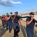 A group of U.S. Coast Guard personnel in navy blue uniforms are carrying a large black hose along a pier or dock area. The team appears to be conducting a training exercise or operational drill, working cooperatively to transport the heavy equipment. The setting appears to be a waterfront facility with a large vessel visible in the background.
