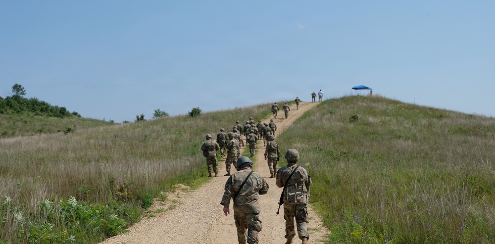 A column of military personnel in camouflage uniforms marches along a narrow dirt path ascending a grassy hill under a clear blue sky. The soldiers appear to be on a training exercise or ruck march, moving in a loose formation toward the hilltop where a blue canopy tent and a few individuals are visible. The setting appears to be a military training area with open grassland terrain.
