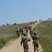 A column of military personnel in camouflage uniforms marches along a narrow dirt path ascending a grassy hill under a clear blue sky. The soldiers appear to be on a training exercise or ruck march, moving in a loose formation toward the hilltop where a blue canopy tent and a few individuals are visible. The setting appears to be a military training area with open grassland terrain.