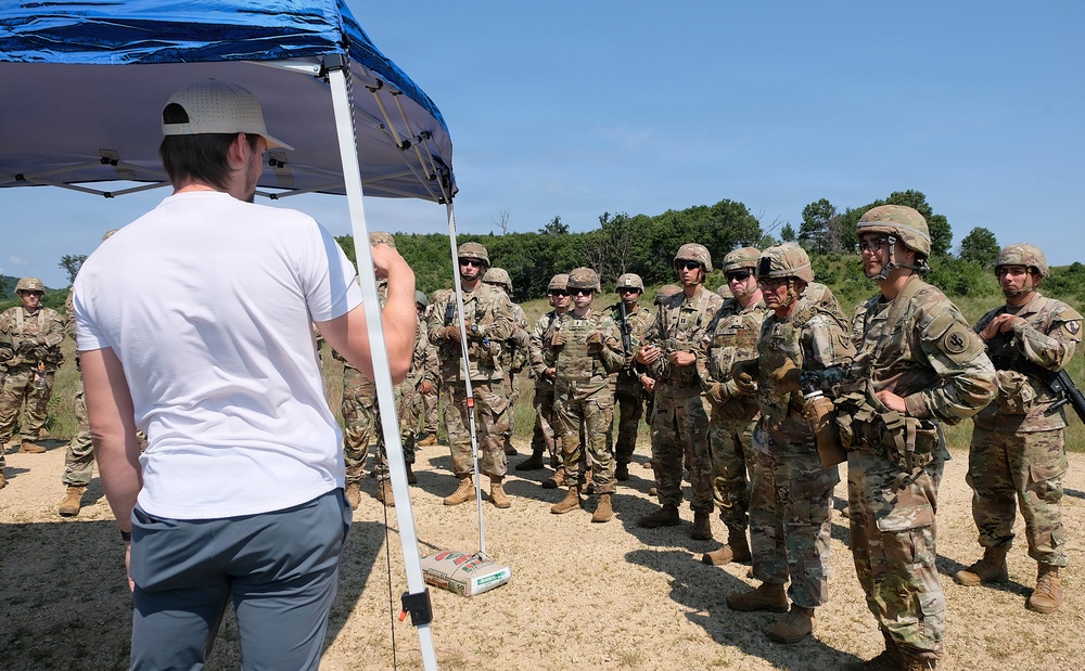 A civilian instructor in a white t-shirt and baseball cap addresses a group of military personnel in full combat gear and camouflage uniforms at an outdoor training site. The soldiers are gathered in a semicircle listening to the briefing under a blue pop-up canopy tent. The setting appears to be a military training range or exercise area with open terrain and tree line in the background.