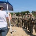 A civilian instructor in a white t-shirt and baseball cap addresses a group of military personnel in full combat gear and camouflage uniforms at an outdoor training site. The soldiers are gathered in a semicircle listening to the briefing under a blue pop-up canopy tent. The setting appears to be a military training range or exercise area with open terrain and tree line in the background.