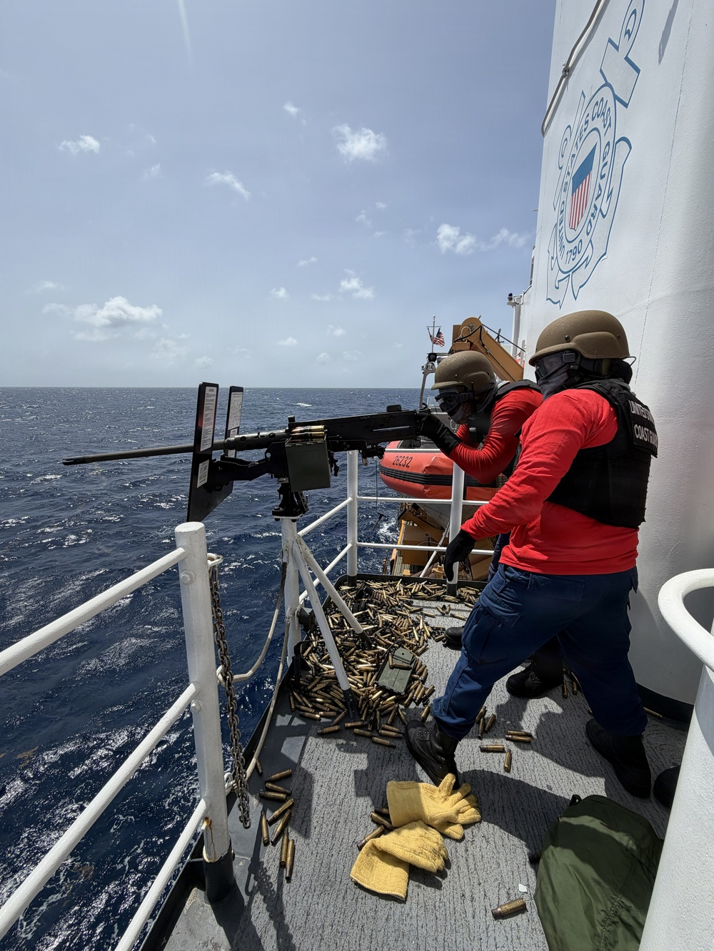 Two United States Coast Guard personnel are conducting a live-fire exercise on the deck of a Coast Guard vessel at sea. One operator is firing a mounted machine gun (appears to be an M2 .50 caliber) while a second crew member assists. A large number of spent brass casings are scattered across the deck, indicating sustained firing.