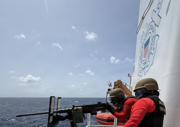 Two United States Coast Guard personnel are conducting a live-fire exercise on the deck of a Coast Guard vessel at sea. One operator is firing a mounted machine gun (appears to be an M2 .50 caliber) while a second crew member assists. A large number of spent brass casings are scattered across the deck, indicating sustained firing.