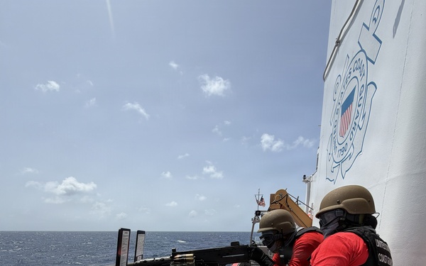Two United States Coast Guard personnel are conducting a live-fire exercise on the deck of a Coast Guard vessel at sea. One operator is firing a mounted machine gun (appears to be an M2 .50 caliber) while a second crew member assists. A large number of spent brass casings are scattered across the deck, indicating sustained firing.