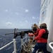 Two United States Coast Guard personnel are conducting a live-fire exercise on the deck of a Coast Guard vessel at sea. One operator is firing a mounted machine gun (appears to be an M2 .50 caliber) while a second crew member assists. A large number of spent brass casings are scattered across the deck, indicating sustained firing.