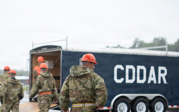 Military personnel in camouflage uniforms and orange hard hats are working around a large dark blue trailer marked 'CDDAR' on a wet tarmac or airfield surface. CDDAR stands for Crashed, Damaged, or Disabled Aircraft Recovery, indicating this is likely a training exercise or operational deployment of aircraft recovery equipment. The overcast, rainy conditions suggest field operations under adverse weather.