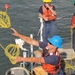 The image shows crew members aboard a vessel engaged in a line-throwing exercise or mooring operation on open water. A person wearing a blue hard hat and orange life vest is actively throwing a coiled yellow heaving line with a red monkey's fist knot at its end. A second crew member in a yellow hard hat and orange life vest stands in the background communicating via radio.