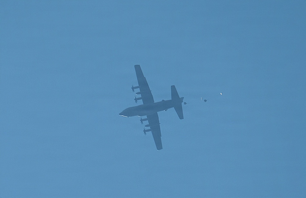 A large military transport aircraft, consistent with a C-130 Hercules configuration, is photographed from below against a clear blue sky. The aircraft appears to be conducting an airdrop operation, with a small object and what appears to be a partially deployed parachute visible to the right of the aircraft. The image is taken at significant altitude, rendering the aircraft as a dark silhouette.
