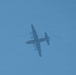 A large military transport aircraft, consistent with a C-130 Hercules configuration, is photographed from below against a clear blue sky. The aircraft appears to be conducting an airdrop operation, with a small object and what appears to be a partially deployed parachute visible to the right of the aircraft. The image is taken at significant altitude, rendering the aircraft as a dark silhouette.