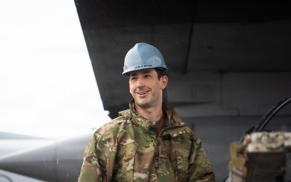 A military service member in OCP (Operational Camouflage Pattern) uniform is working on a flight line near a military aircraft. The individual is wearing a blue hard hat and an orange high-visibility safety belt, holding what appears to be a fuel hose or ground support cable. A US Air Force aircraft with a visible star-and-bar insignia is visible in the background.