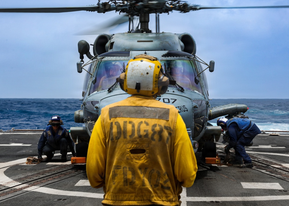 A military helicopter, numbered 707, is on the flight deck of a U.S. Navy destroyer with rotors spinning. Deck crew members in colored jerseys are performing pre-flight or post-landing procedures around the aircraft. The scene takes place at sea under overcast skies with visible ocean swells in the background.