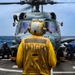 A military helicopter, numbered 707, is on the flight deck of a U.S. Navy destroyer with rotors spinning. Deck crew members in colored jerseys are performing pre-flight or post-landing procedures around the aircraft. The scene takes place at sea under overcast skies with visible ocean swells in the background.