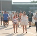 A group of people are walking toward the camera in what appears to be a military homecoming event, likely at a base or port facility. Two young women in the foreground are carrying handmade welcome home signs, one reading 'Welcome Home Petereit' and the other 'Always Worth the Wait, Welcome Home [name] Herrington' with an anchor symbol. The crowd includes families with children, suggesting they are gathering to greet returning service members.