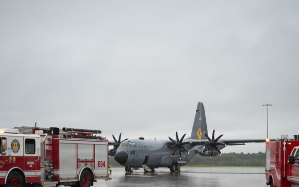 A wet airport tarmac scene showing two red emergency fire vehicles flanking a U.S. Air Force C-130 turboprop military transport aircraft. The scene appears to be a ceremonial or precautionary escort on a rainy day at a military airfield. The reflective wet pavement and overcast sky create a moody, low-light atmosphere.