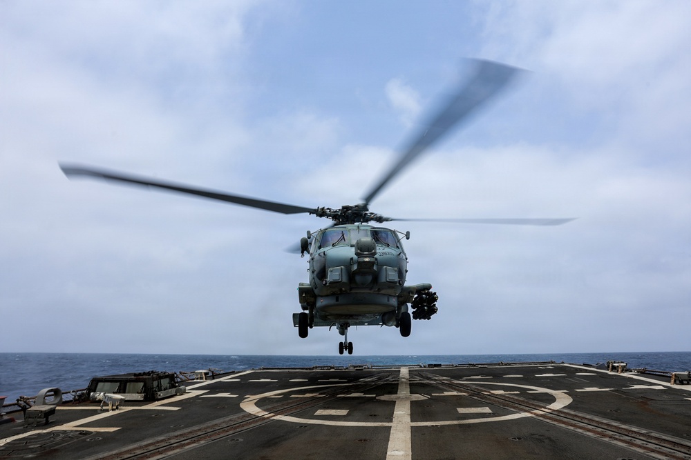 A military helicopter is captured in a hover or low-altitude flight directly above a naval vessel's flight deck at sea. The rotor blades are visibly motion-blurred, indicating active rotation at high speed. The scene depicts a shipboard aviation operation, likely a takeoff or landing evolution.