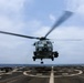 A military helicopter is captured in a hover or low-altitude flight directly above a naval vessel's flight deck at sea. The rotor blades are visibly motion-blurred, indicating active rotation at high speed. The scene depicts a shipboard aviation operation, likely a takeoff or landing evolution.