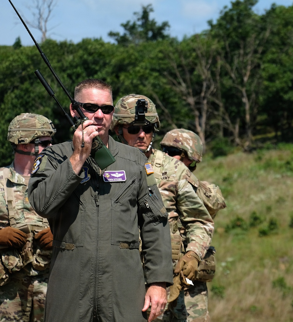 A military personnel wearing an olive drab flight suit is using a handheld radio with an extended antenna in an outdoor field setting. He is accompanied by several soldiers in Army Combat Uniform (ACU) with helmets and tactical gear. The scene appears to be a joint military exercise or training operation involving Air Force and Army personnel.