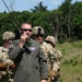 A military personnel wearing an olive drab flight suit is using a handheld radio with an extended antenna in an outdoor field setting. He is accompanied by several soldiers in Army Combat Uniform (ACU) with helmets and tactical gear. The scene appears to be a joint military exercise or training operation involving Air Force and Army personnel.