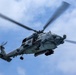 A U.S. Navy military helicopter, marked with the number '707' and the word 'NAVY,' is photographed from below while in flight against a partly cloudy blue sky. The helicopter appears to be an MH-60R Seahawk, identifiable by its distinctive nose-mounted sensor dome and overall configuration. The main rotor blades show motion blur, indicating active flight.