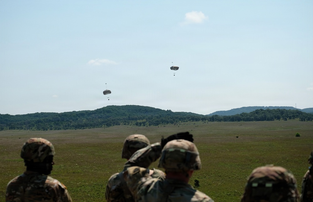 Military personnel in camouflage uniforms observe two parachute drops over a large open field. The parachutes appear to be carrying cargo or equipment loads descending toward a drop zone. The scene takes place in a rural, hilly landscape under partly cloudy skies.
