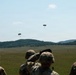 Military personnel in camouflage uniforms observe two parachute drops over a large open field. The parachutes appear to be carrying cargo or equipment loads descending toward a drop zone. The scene takes place in a rural, hilly landscape under partly cloudy skies.