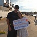 A U.S. Coast Guard servicemember identified as FN Herrington is reunited with a woman, likely a family member or partner, at a naval pier following a deployment. The woman holds a handmade welcome home sign reading 'Always Worth the Wait Welcome Home FN Herrington' with a heart and anchor symbol. The scene takes place dockside with a large Coast Guard vessel visible in the background.
