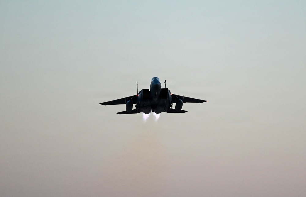 A twin-engine military jet aircraft is photographed head-on in silhouette against a hazy, muted sky. The aircraft is in flight with both afterburners visibly ignited, producing bright white exhaust flames beneath the fuselage. The low-light, backlit conditions render the aircraft almost entirely in dark silhouette.