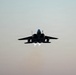 A twin-engine military jet aircraft is photographed head-on in silhouette against a hazy, muted sky. The aircraft is in flight with both afterburners visibly ignited, producing bright white exhaust flames beneath the fuselage. The low-light, backlit conditions render the aircraft almost entirely in dark silhouette.