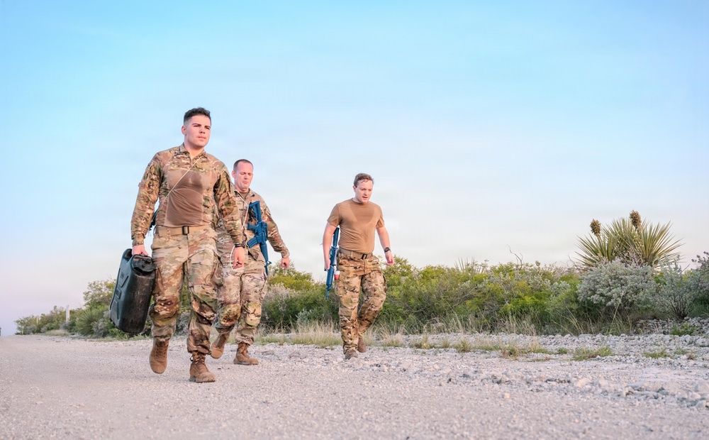 Three military personnel in combat uniforms are walking along a gravel path in an arid, desert-like environment. Two of the individuals are wearing full camouflage uniforms while the third wears a tan t-shirt with camouflage pants. At least two of them are carrying blue training rifles, suggesting a training exercise rather than a combat deployment.