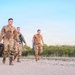 Three military personnel in combat uniforms are walking along a gravel path in an arid, desert-like environment. Two of the individuals are wearing full camouflage uniforms while the third wears a tan t-shirt with camouflage pants. At least two of them are carrying blue training rifles, suggesting a training exercise rather than a combat deployment.