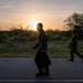 Two individuals in military-style camouflage uniforms are walking along a paved road at sunset or sunrise. The backlit scene creates a dramatic silhouette effect, with warm golden light emanating from behind the lead figure. The surrounding landscape features scrubland vegetation and sparse trees typical of a semi-arid environment.