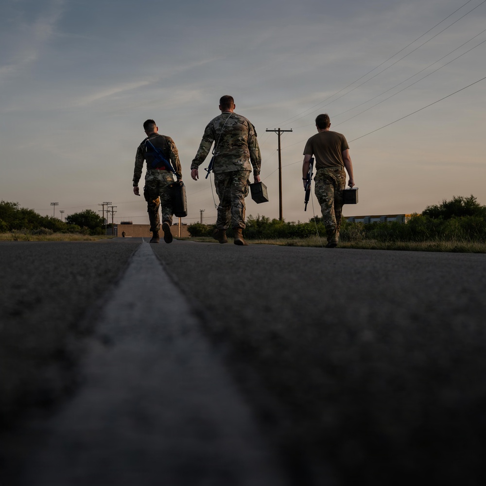 Three military personnel in camouflage uniforms are walking away from the camera along a paved road at dusk or dawn. They are carrying equipment cases and appear to be moving toward a destination in a semi-rural or base environment. The low-angle shot creates a dramatic silhouette effect against a partly cloudy sky.