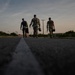 Three military personnel in camouflage uniforms are walking away from the camera along a paved road at dusk or dawn. They are carrying equipment cases and appear to be moving toward a destination in a semi-rural or base environment. The low-angle shot creates a dramatic silhouette effect against a partly cloudy sky.