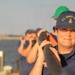 A U.S. Coast Guard member in the foreground wearing a navy blue 'U.S. Coast Guard' cap and uniform is holding onto a railing or boom, smiling slightly. Behind her, multiple personnel in similar uniforms are using binoculars to scan the water. The scene appears to take place on or near a vessel with a body of water visible in the background.