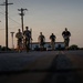 Five individuals in military-style camouflage uniforms are moving at a jog or quick walk along a road at dusk or dawn. They are carrying various equipment including what appear to be weapons and supply cases. A street sign reading 'Indiana Blvd' and utility poles are visible in the background near a portable building.