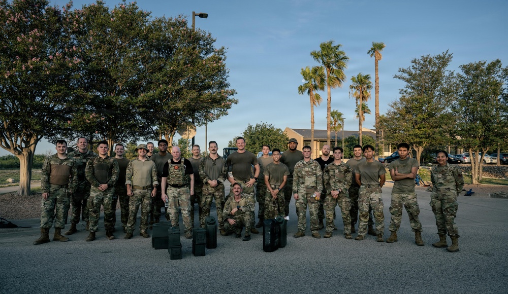 A large group of approximately 25 military personnel posing for a group photograph outdoors, likely on a military installation. Most individuals are wearing MultiCam/OCP camouflage uniforms, while a few wear solid olive/dark colored shirts. Several ammunition cans and equipment cases are visible on the ground in front of the group.